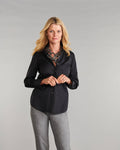 A blonde woman wearing a black long-sleeve button-up blouse paired with gray heather pants, standing against a neutral gray studio background.